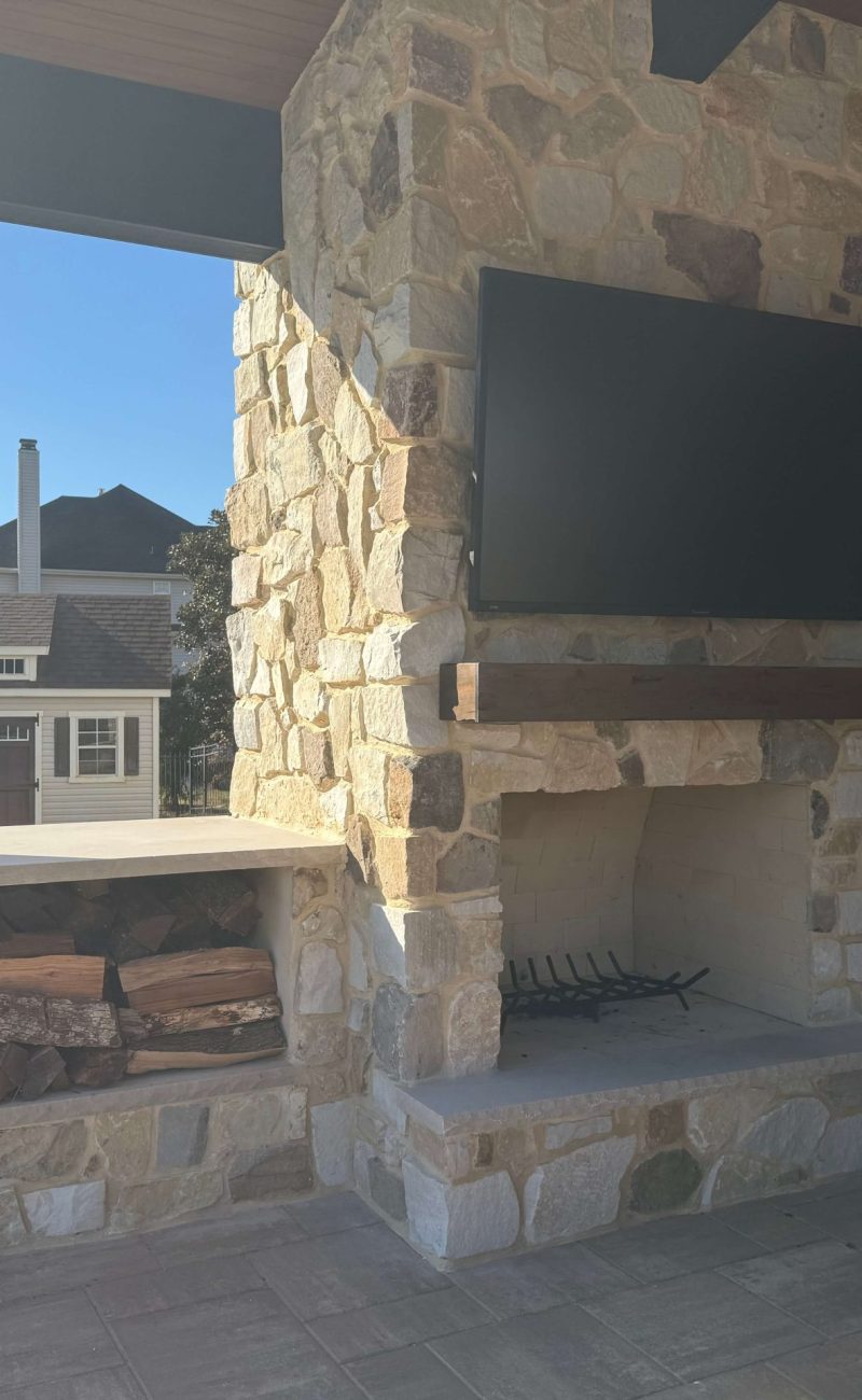 Outdoor stone fireplace with mounted TV and built-in firewood storage on covered patio in South Jersey